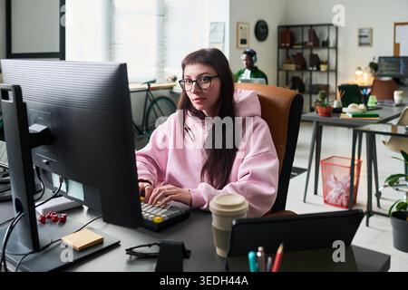 Young adult Caucasian woman working at desktop computer in modern office, typing and focusing on digital tasks. Useful for remote work, startup, productivity, coding themes Stock Photo