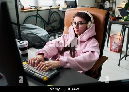 Young adult Caucasian woman wearing headphones and typing at computer in home office, working remotely. Ideal for remote work, gaming, digital lifestyle, productivity marketing Stock Photo