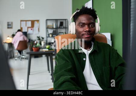 Portrait of young Black man wearing headphones, working at computer in office, looking at camera and supporting remote collaboration. Ideal for hybrid work or team communication campaigns Stock Photo