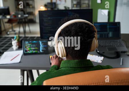 Rear view of young Black man wearing headphones, coding across multiple screens at desk, representing software development and remote work. Ideal for tech careers or IT services Stock Photo