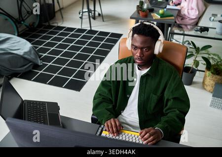 Young African American man working at desktop computer with headphones in home office, typing and focusing on task. Ideal for remote work, productivity, digital career content Stock Photo