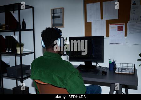 Young Black man wearing headphones working at desktop computer, coding in home office and reviewing data charts. Ideal for remote work, software development, productivity content Stock Photo