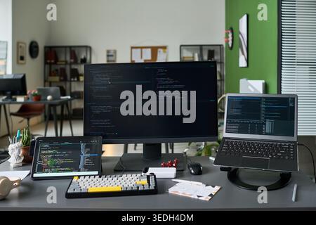 Wide shot of software development workspace displaying code on desktop monitor, laptop, tablet. Supporting programming, app creation, cybersecurity, tech marketing content Stock Photo