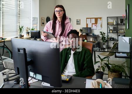 Portrait of diverse young adult coworkers collaborating with desktop computer and tablet in office, representing teamwork and digital workflow. Ideal for business communication and startup marketing Stock Photo