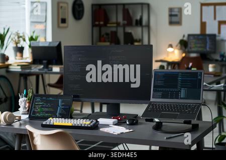Wide shot of developer workstation displaying code on desktop monitor, laptop, tablet in modern office. Supporting software development, remote work, cybersecurity marketing Stock Photo