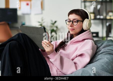 Young adult Caucasian woman wearing headphones and using tablet while relaxing at home, streaming online content and mobile learning. Ideal for remote lifestyle, digital media, e learning Stock Photo