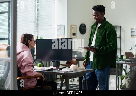 Young Black man discussing project with Caucasian young adult woman at office desk, holding tablet and reviewing work. Ideal for teamwork, startup, workplace communication themes Stock Photo