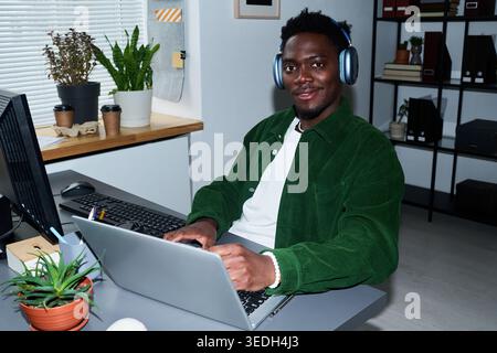 Portrait of young Black man working on laptop at desk, wearing headphones and looking at camera in office. Ideal for remote work, productivity, tech support, corporate communication Stock Photo