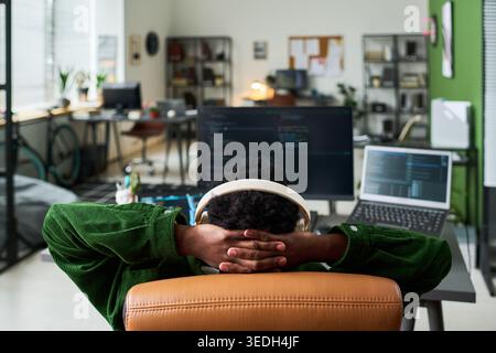 Young African American man reclining at coding workstation, wearing headphones and resting after programming session. Ideal for remote work, developer burnout, tech productivity Stock Photo