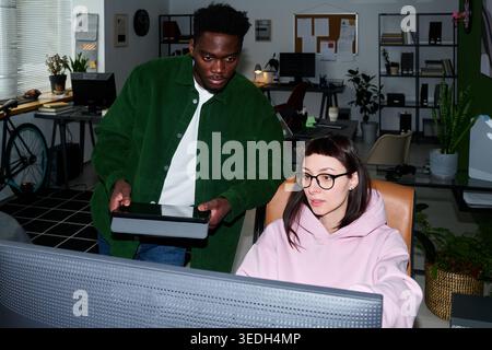 African American young man showing tablet to Caucasian young woman working at desktop monitor in office, collaborating on project. Ideal for teamwork, startup, workplace communication Stock Photo