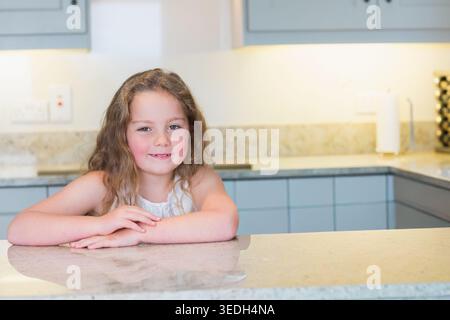 Female child leaning on stone counter in home kitchen, wearing white sleeveless top, near wine rack. Portrait, modern, bright, minimal, neutral, grani Stock Photo