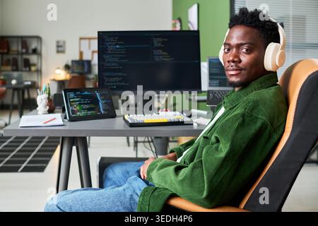 Portrait of young Black man sitting at workstation, coding on desktop monitor and tablet while wearing headphones. Ideal for software development, tech careers, remote work marketing Stock Photo