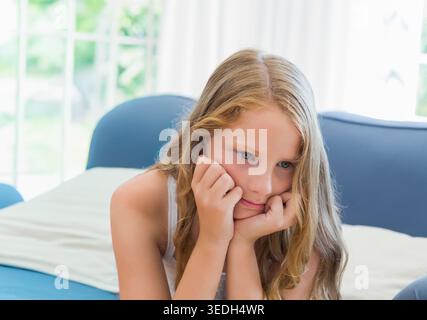 Female child lying on bed in white tank top, resting chin on hands, gazing out window. Girl, contemplative, sunlit, bedroom, sofa, greenery, blue Stock Photo