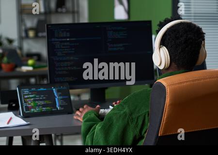 Rear view of young Black man coding at desktop computer and tablet while wearing headphones in home office. Ideal for software development, remote work, tech career content Stock Photo