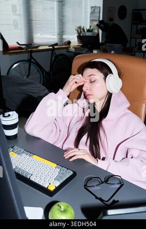 Vertical shot of young adult Caucasian woman wearing headphones, rubbing eyes while sitting at computer desk, showing screen fatigue during remote work. Ideal for burnout or work stress campaigns Stock Photo