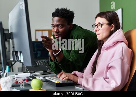 Young adult Black man guiding Caucasian woman while reviewing code on desktop monitor in office. Depicting teamwork and software development for tech hiring or training campaigns Stock Photo