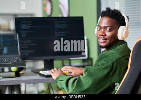 Portrait of young Black man coding at desktop workstation while wearing headphones and typing on keyboard, representing software development and tech careers. Ideal for IT hiring, programming courses Stock Photo