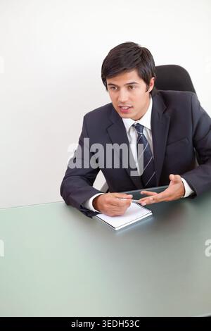 Adult man in suit and tie sitting at glass desk, holding pen over spiral notepad, gesturing. Executive, professional, office, notebook, discussion, co Stock Photo