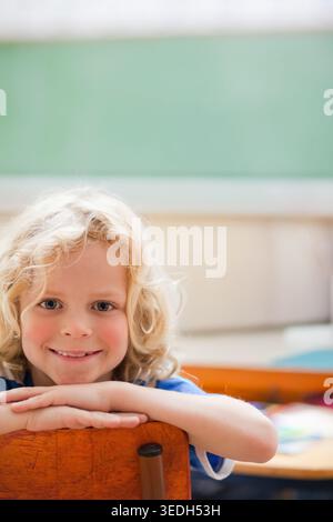 Child boy leaning on wooden chair, smiling, wearing blue short-sleeved shirt near green chalkboard. Classroom, student, desks, learning, daylight, sof Stock Photo