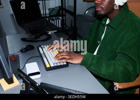 Close up of young Black man typing on keyboard at desk, wearing headphones and working on computer in home office. Ideal for remote work, productivity, tech lifestyle marketing Stock Photo