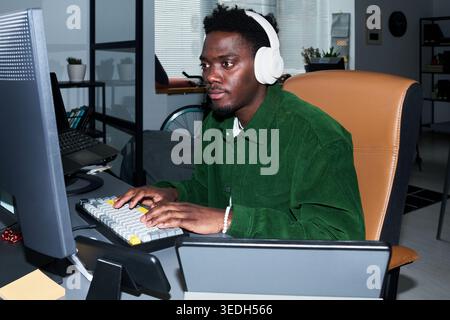 Young African American man wearing headphones working at desktop computer and typing at home office, supporting remote work, online productivity, digital business content Stock Photo