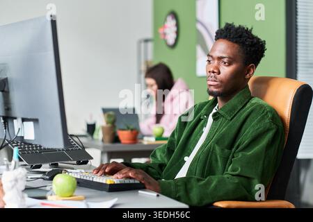 Medium shot of African American young adult man working at desktop computer in office, focusing on task and productivity. Suitable for workplace, career growth, team workflow marketing Stock Photo