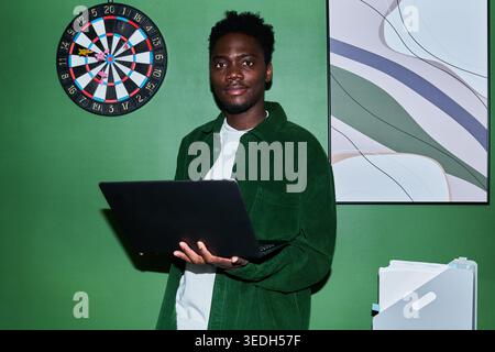 Portrait of young Black man holding laptop and looking at camera beside dartboard, suggesting focus and productivity. Useful for remote work, goals, planning, business content Stock Photo