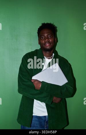 Vertical portrait of young Black man holding laptop and looking at camera against green wall, presenting remote work and digital professional identity. Useful for career, tech  Stock Photo