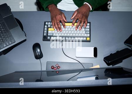Overhead shot of Black young adult man typing on keyboard at workstation with laptop and smartphone, showing digital productivity. Ideal for remote work, tech workflow, office use Stock Photo