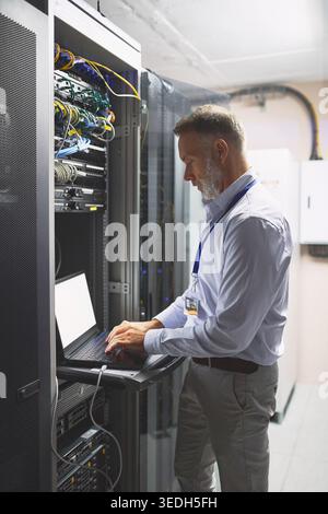 Engineer, typing and man with laptop screen in server room, online or system maintenance on website. Serious, mature person and network management Stock Photo