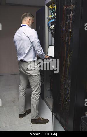 Man, typing and programming with laptop in server room for research, coding and cybersecurity. Mature person, mainframe and tech with software update Stock Photo