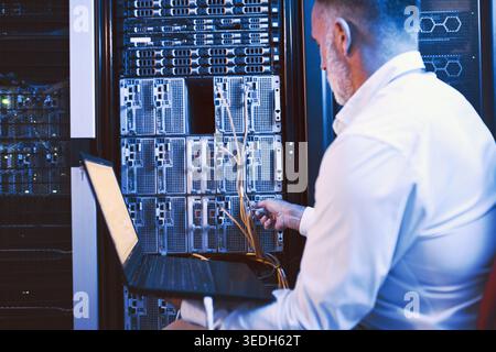Man, servers and cables with laptop in data center for research, coding and cybersecurity. Mature person, mainframe or technology for software update Stock Photo