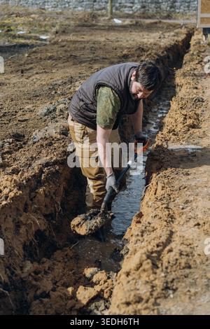 A man digs a drainage trench on his countryside property, working outdoors with a shovel. Vertical photo Stock Photo