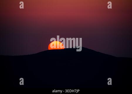 Half sun rests on the line of a dark hill glowing against a deep magenta sky. Clean shapes and intense color make a powerful graphic moment at the clo Stock Photo