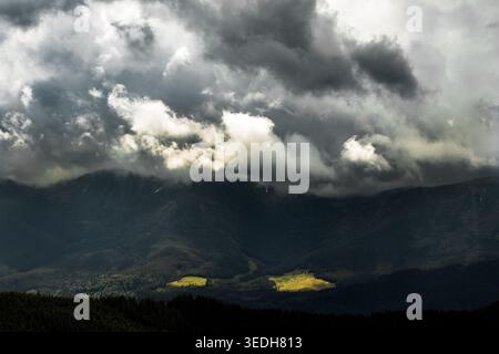 Storm clouds churn above a vast mountain basin while rare sunbeams paint glowing patches on distant meadows a powerful contrast of darkness and hope a Stock Photo