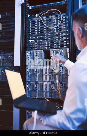 Man, mainframe and cables with laptop in server room for research, coding and cybersecurity. Mature person, database and tech for software update Stock Photo