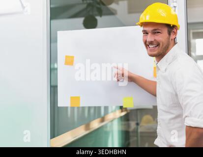Man wearing yellow hard hat pointing at blank poster with sticky notes on glass, copy space. Engineer, business, modern, construction, office, collabo Stock Photo