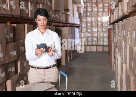 Female wearing white shirt khakis using scanner checking boxes in storage aisle by shelves cart. Warehouse, inventory, logistics, organized, bright, t Stock Photo
