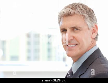 Senior man posing for headshot at office window in dark suit and tie, copy space. Portrait, executive, backlit, cityscape, urban, professional, confid Stock Photo