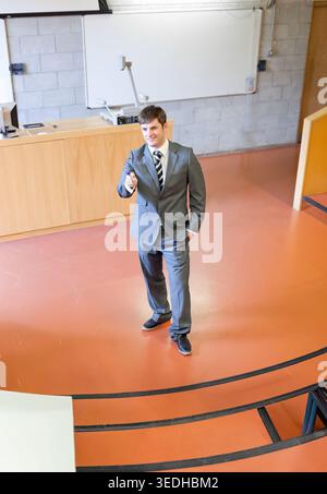 Man in grey suit standing and presenting with remote on lecture theatre stage near whiteboard. Speaker, classroom, professional, talk, projection, scr Stock Photo