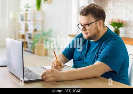 Overweight man taking notes while studying online at home with laptop on desk Stock Photo