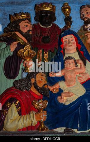 Adoration of the Magi: three wise men or kings from the east present Christ Child with gifts of gold, frankincense and myrrh in charming late 17th or early 18th century polychrome bas-relief on wooden altarpiece in former Augustinian abbey of Our Lady or Notre-Dame at Daoulas, North Finistere, Brittany, France.  Painted rustic Baroque sculpture typical of local Breton churches. Stock Photo