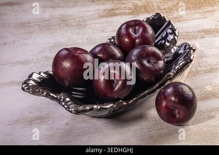 Close-up still life, ripe deep purple plums in decorative shiny dark bowl. One plum rests beside bowl on textured light rustic surface Stock Photo