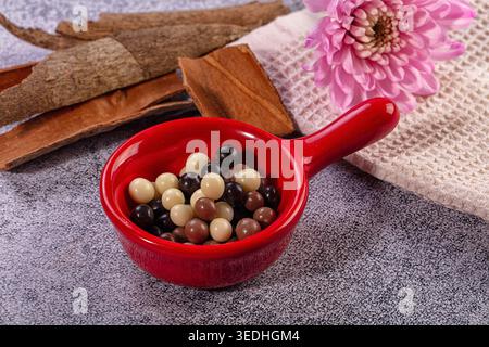 Close up view small dark, milk, white chocolate balls vibrant red ceramic bowl. Cinnamon sticks, pink flower, waffle-weave towel arranged on speckled Stock Photo