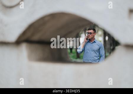 Businessman talking on phone outdoors framed by a stone sculpture opening in urban park Stock Photo