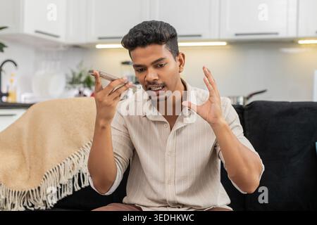 Indian man at home records voice message on smartphone speaks clear checks mic level before reply. Arabian guy on sofa talks in audio chat uses gesture for emphasis sends message and ends recording Stock Photo