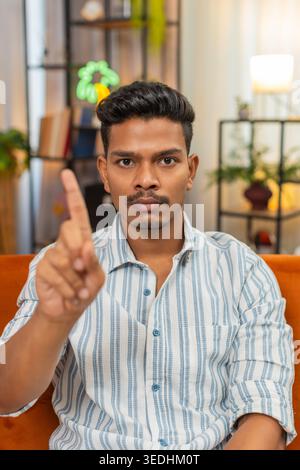 Indian man at home waves finger no no to camera and shows refusal and disagreement with stern mood. Arabian guy on sofa shakes head denies request points finger and shows unhappy sad reject reaction Stock Photo