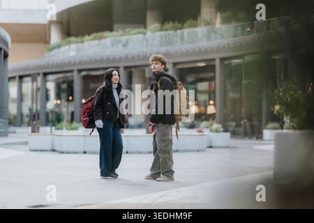 High school friends standing with backpacks in an urban plaza talking and smiling casually Stock Photo