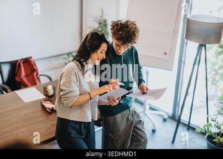 Two students collaborating over documents in a bright modern classroom while smiling and discussing Stock Photo