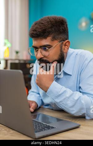 Indian man at home types on laptop stops rubs chin thinking over vital idea creative future plan. Arabian guy at table gazes screen focused eyes show deep concentration solving problem quickly today. Stock Photo